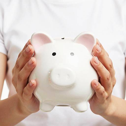 Close-up photograph of a person's hands holding a white, porcelain piggy bank with pink-tinted ears and a small black hole in the center