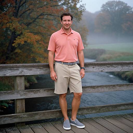 Photograph of a smiling man with short brown hair, wearing a pink polo, beige shorts, and gray sneakers, standing on a wooden bridge with a