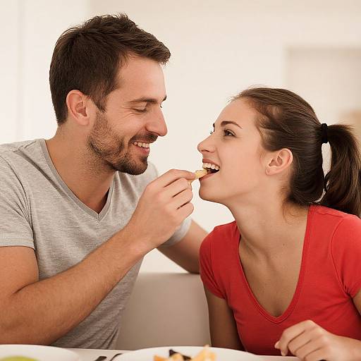 Photograph of smiling, dark-haired man in gray shirt feeding bite to brown-haired woman in red shirt at white table.