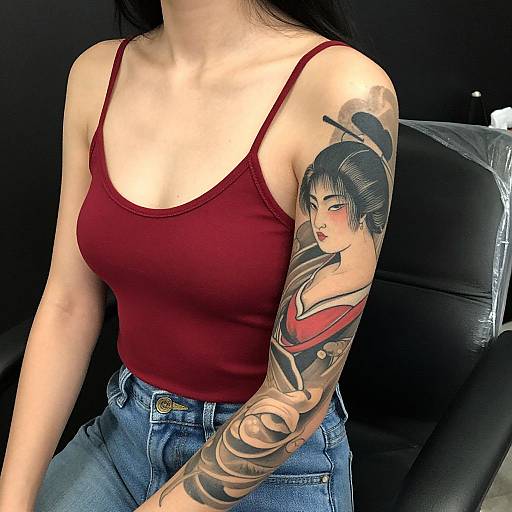 Asian woman in red tank top and blue jeans, revealing detailed anime-style tattoo sleeve of a female warrior on her right arm. Photographic image.