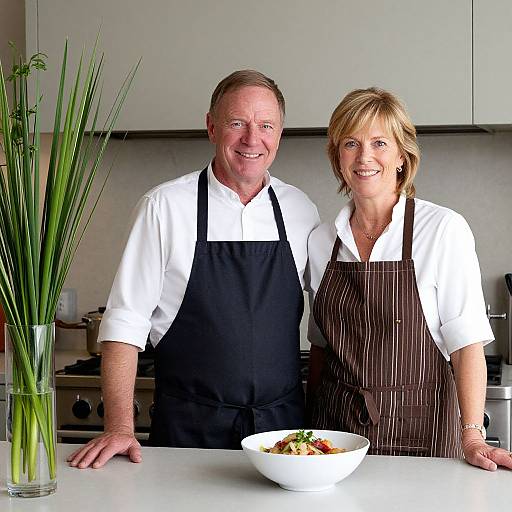 Photograph of smiling middle-aged Caucasian chef and waitress in white shirts and black/brown striped aprons, standing in modern kitchen with white countertops and tall