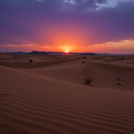 Photograph of a vivid desert sunset with a bright orange sun, purple and blue sky, and rippled sand dunes in the foreground.
