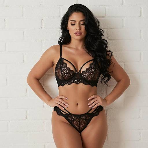 Photograph of a curvy woman with long black hair, wearing black lace lingerie, standing against a white brick wall.