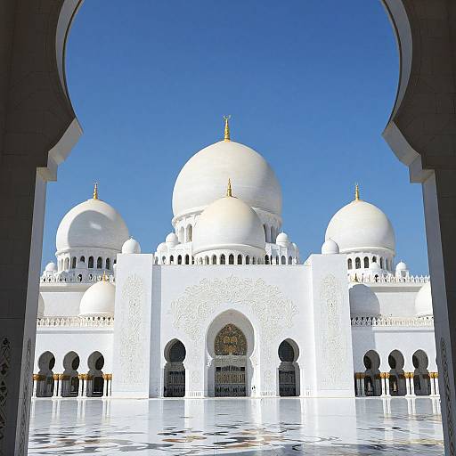 Photograph of a white, domed mosque with intricate patterns, viewed through an archway, against a clear blue sky. Reflective marble courtyard in