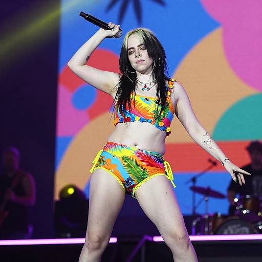 Photograph of a pale, black-haired woman in a colorful, tie-dye bikini, holding a microphone, performing on a brightly lit stage with abstract
