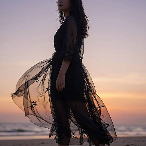 Silhouetted woman in black sheer dress with floral patterns, standing against a sunset beach backdrop, waves in the distance. Photograph.