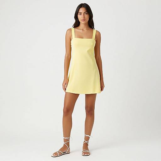 Photograph of a young woman with dark hair wearing a sleeveless, yellow mini dress and white strappy sandals, standing against a plain white background.