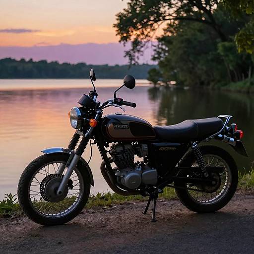 Photograph of a black Royal Enfield motorcycle parked on a dirt path by a serene lake at sunset, with trees and a colorful sky in the background