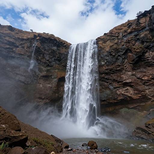 Photograph of a powerful waterfall cascading down rugged, rocky cliffs into a misty pool, with a bright blue sky overhead.