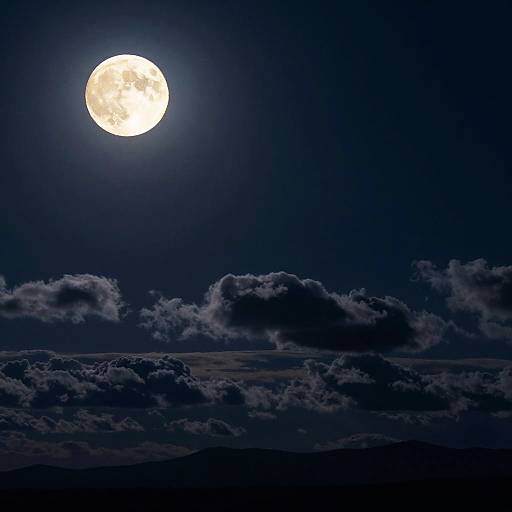 Photograph of a bright full moon in a dark blue night sky, with fluffy clouds below and silhouetted mountain peaks.