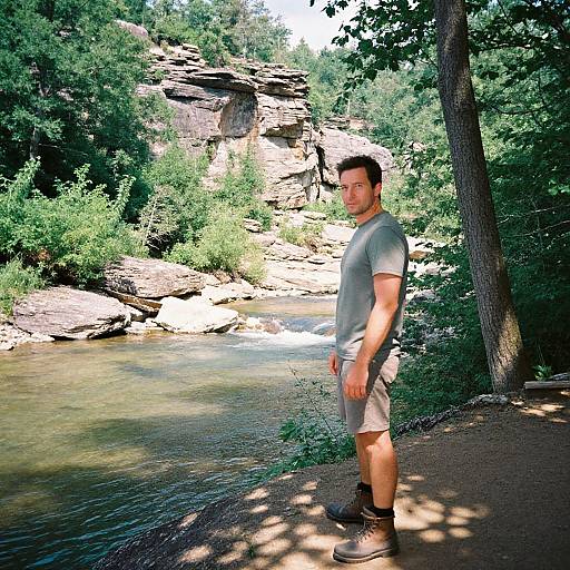 Photograph of a muscular man with short brown hair, wearing a gray t-shirt, gray shorts, and black boots, standing by a rocky stream in