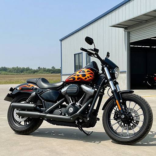 Photograph of a black motorcycle with orange flame decals parked in front of a white metal garage under a clear blue sky.