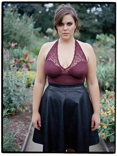 Photograph of a curvy woman with light skin and brown hair in a maroon lace halter top and black skirt, standing in a lush garden