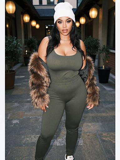 Photograph of a curvy woman with long black hair, wearing a white beanie, green bodysuit, and brown fur sleeves, standing on