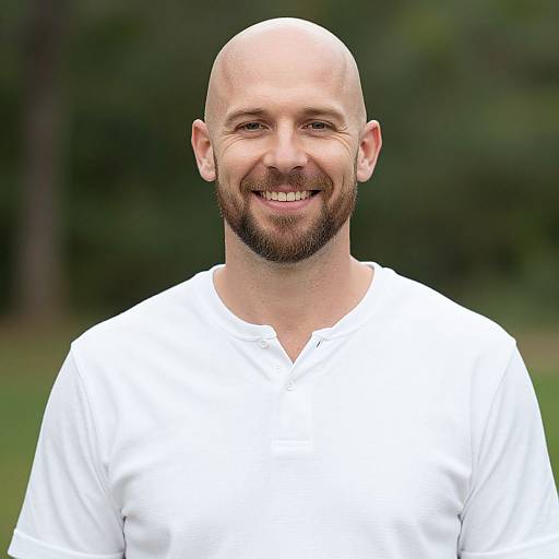 Photograph of a bald, bearded man with a smiling face, wearing a white, V-neck shirt, against a blurred green outdoor background.