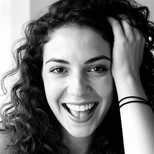 Black-and-white photograph of a smiling woman with curly hair, bright eyes, and hand in hair, wearing a black wristband.