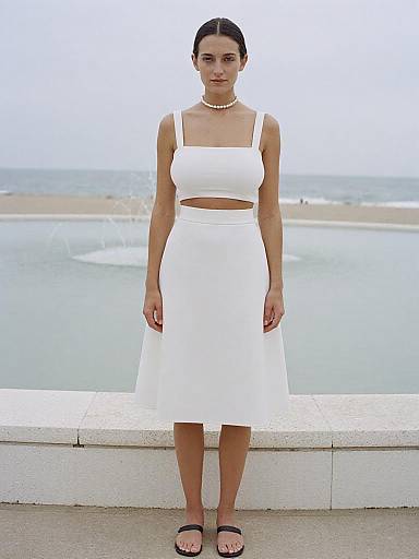 Photograph of a woman with dark hair in a white crop top and skirt, black sandals, pearl necklace, standing by the sea.
