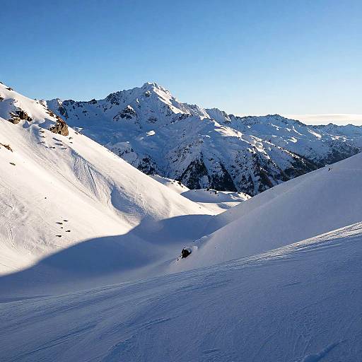Photograph of a snowy mountain landscape with a single skier in a small valley, under a clear blue sky.