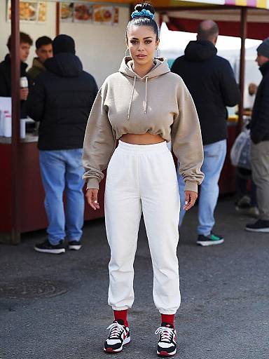 Photograph of a woman with blue hair in a bun, wearing a beige hoodie, white pants, red socks, and black sneakers, standing in an