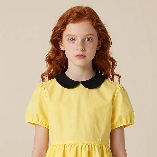 Photograph of a young girl with fair skin and long red curly hair, wearing a bright yellow dress with a black Peter Pan collar, against a plain