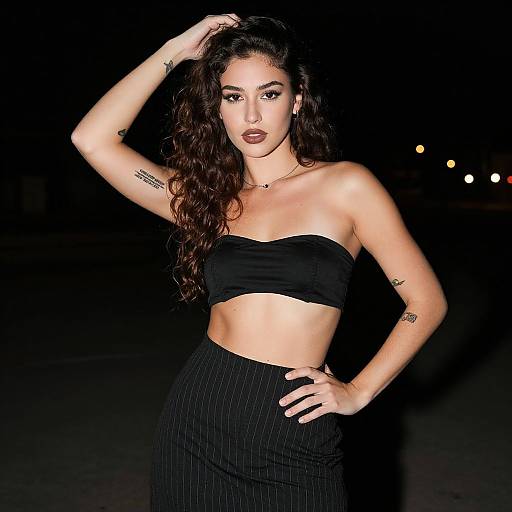 Photograph of a confident, dark-haired woman with tattoos, wearing a black strapless crop top and pinstriped high-waist skirt,