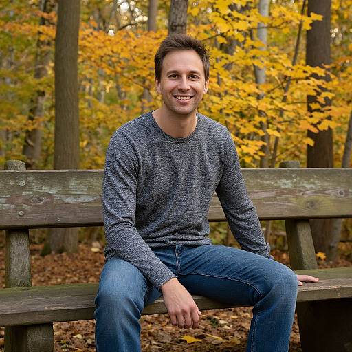 Photograph of a smiling man with short brown hair, wearing a blue speckled sweater and blue jeans, sitting on a wooden bench in a forest