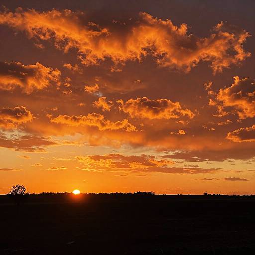 Photograph of a dramatic sunset with vibrant orange and red clouds, a bright sun partially hidden by a dark silhouetted horizon.