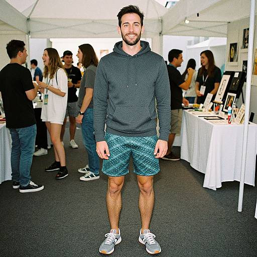 Photograph of a smiling, bearded man in a dark hoodie, teal patterned shorts, and gray sneakers, standing in a busy art exhibition tent