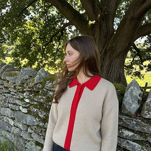 Photograph of a young woman with long brown hair, wearing a beige sweater with a red collar, standing in front of a stone wall and large tree