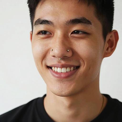 Photograph of a young Asian man with short black hair, smiling, showing three facial piercings: nose and both nostrils, wearing a black