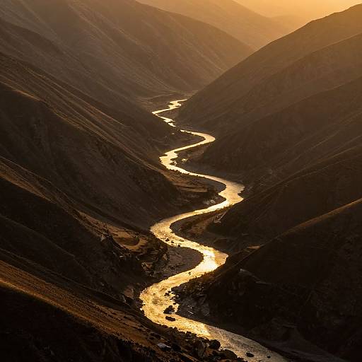 Photograph of a winding, golden river snaking through dark, shadowy mountain valleys during sunset, with warm, orange sunlight casting dramatic light and shadows