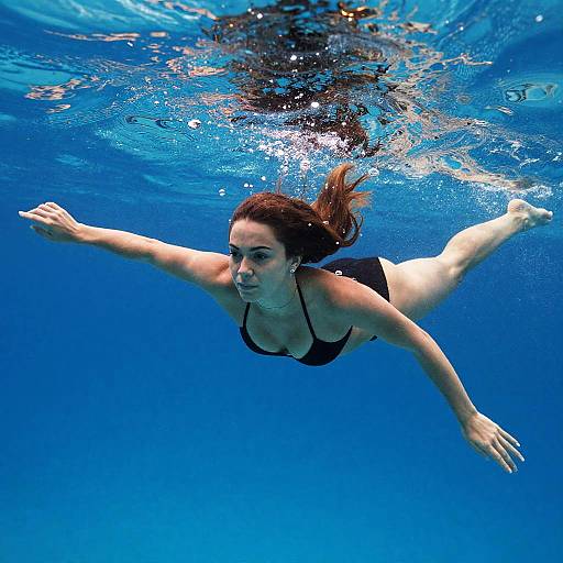 Photograph of a brown-haired woman in a black bikini swimming underwater, arms outstretched, with rippling blue water around her.