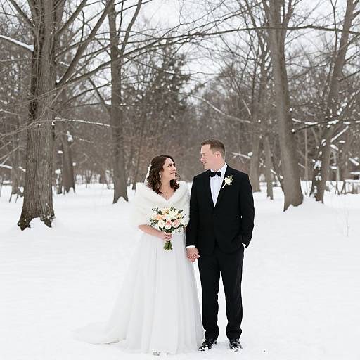 Photograph of a smiling bride in white gown and groom in black tuxedo, holding bouquet, standing in snowy forest with bare trees.