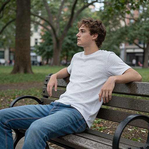 Young man with curly brown hair, wearing a white t-shirt and blue jeans, sits relaxed on a wooden park bench, gazing into the distance.