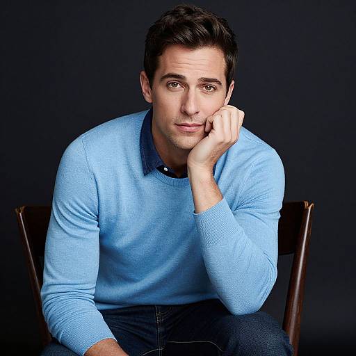 Photograph of a young, dark-haired man with fair skin, wearing a light blue sweater, sitting on a wooden chair against a dark background, resting