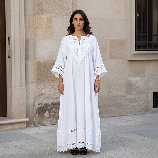 Photograph of a young woman with dark wavy hair wearing a long white embroidered dress, standing against a beige stone building.