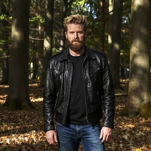 Photograph of a bearded man with blonde hair, wearing a black leather jacket and jeans, standing in a sunlit forest.