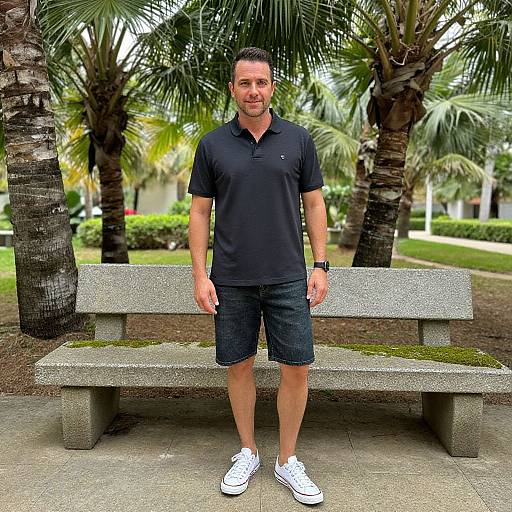 Photograph of a middle-aged man with short dark hair, wearing a black polo, dark shorts, and white sneakers, standing in front of a concrete