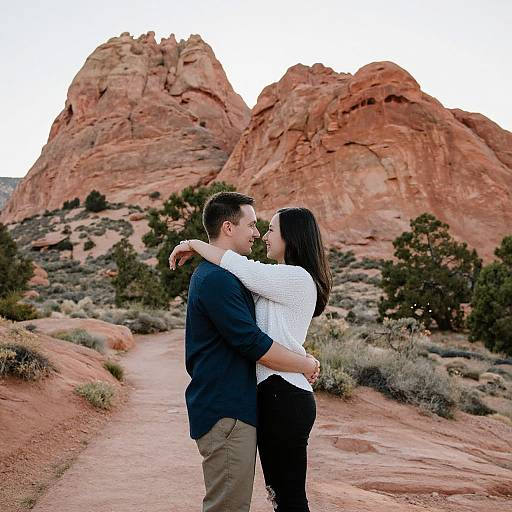 Couple embracing on a desert path with red rock formations in the background, wearing casual clothes, smiling, and standing close. Photographic image.