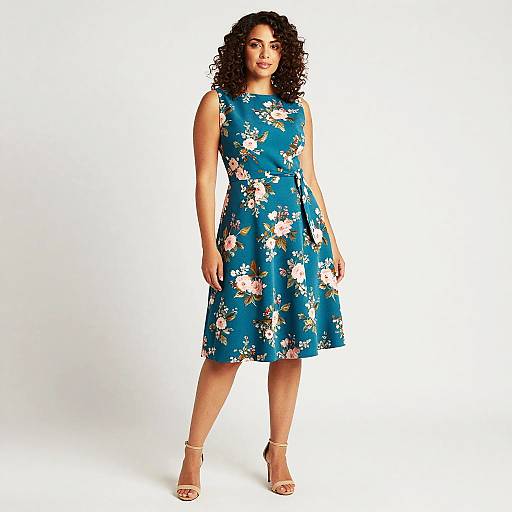 Photograph of a curvy woman with dark curly hair, wearing a blue floral dress and beige heels, standing against a white background.