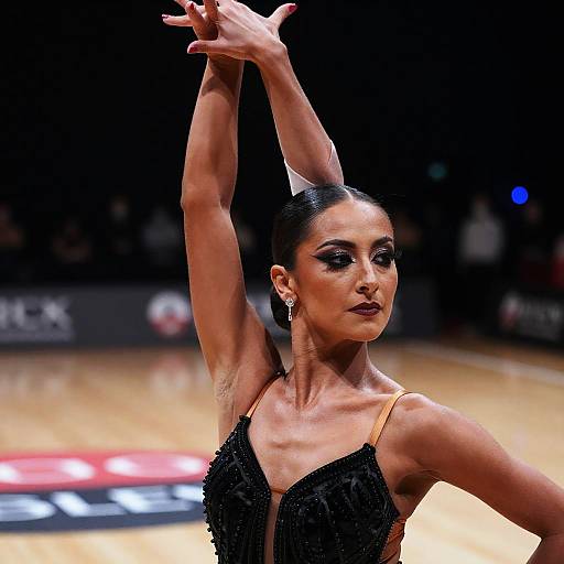 Photograph of a poised, dark-haired ballerina with black, beaded dress, raised arms, and intense gaze, on a brightly lit stage