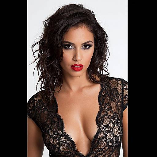Photograph of a woman with dark, wavy hair, red lipstick, and black lace deep V-neck top, against a white background.