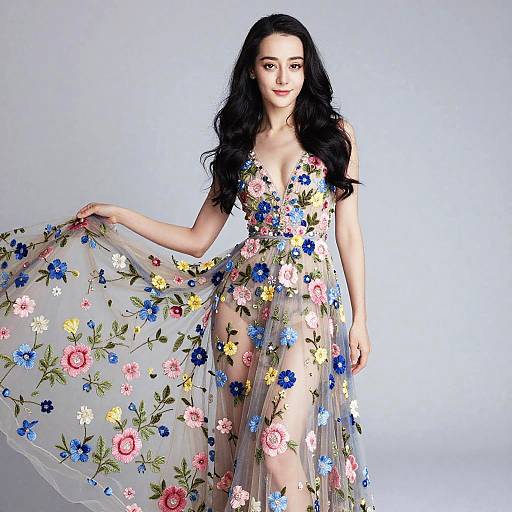 Photograph of a fair-skinned woman with long black hair, wearing a sheer, floral-embellished dress, holding the fabric, against a