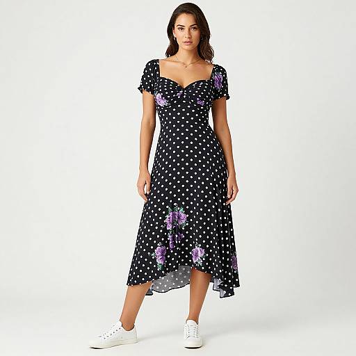 Photograph of a woman with dark hair, wearing a black, polka-dotted dress with purple floral accents, and white sneakers, standing against a