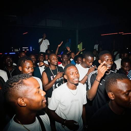 Photograph of a crowded, energetic nighttime party with dark-skinned men in black and white athletic wear, clapping and smiling, illuminated by bright flash