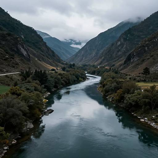 Photograph of a serene, narrow river winding through a lush, mountainous valley with dense greenery, reflecting the cloudy, overcast sky.