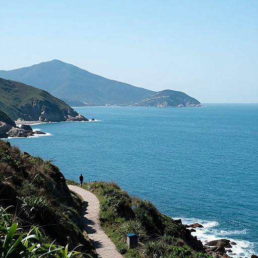 Photograph of a coastal path leading to a blue ocean, with a person walking, green grass, and mountainous cliffs in the background. Clear,