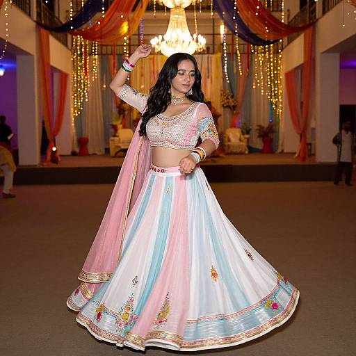 Photograph of a South Asian woman in a white traditional lehenga with pink and gold embroidery, dancing in a brightly lit, decorated hall with hanging lights