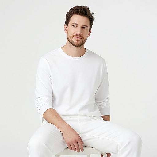 Photograph of a smiling, bearded man with short dark hair, wearing a plain white long-sleeve shirt and pants, sitting against a completely