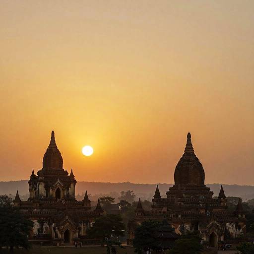 Photograph of Bagan sunrise, showcasing two silhouetted pagodas with pointed roofs against a glowing orange sky, with the sun partially visible